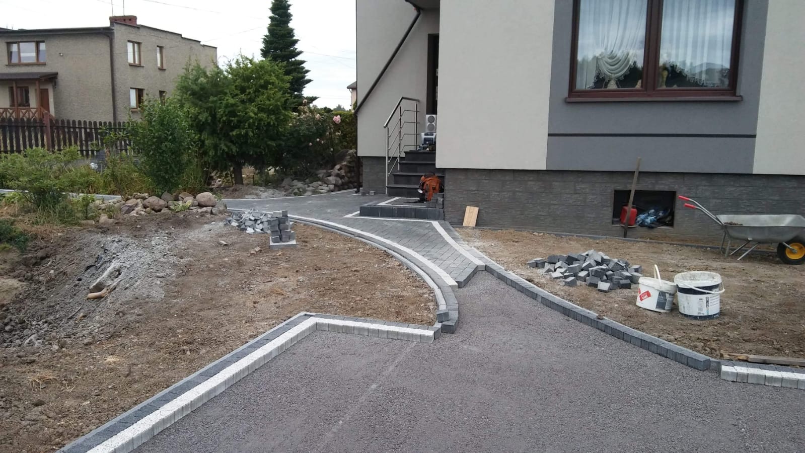 Fragment podjazdu z szarej kostki brukowej z białymi obrzeżami w trakcie układania, widoczne narzędzia i materiały budowlane, dom z oknem w tle.