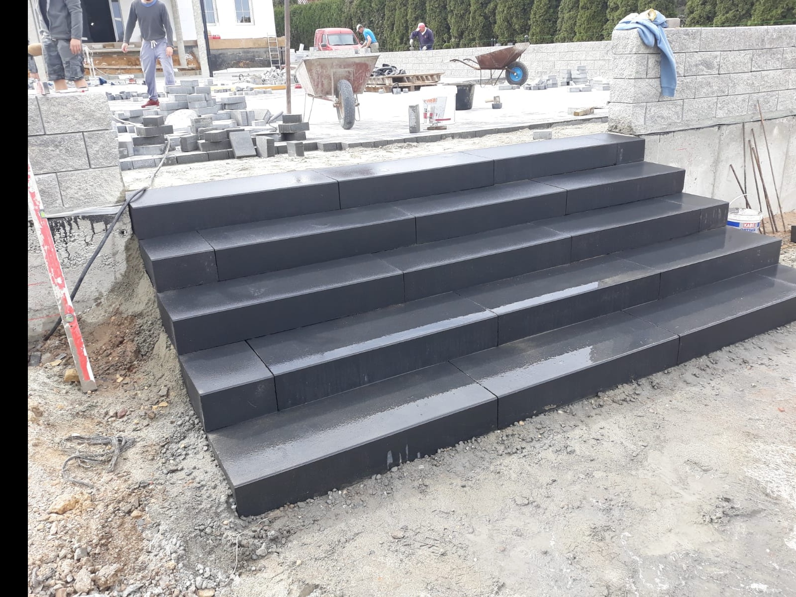 Schody zewnętrzne z ciemnej kostki brukowej, lekko mokre, w trakcie budowy, widoczne narzędzia i materiały budowlane w tle.