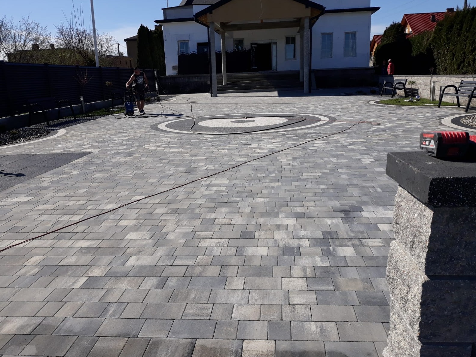 Szara kostka brukowa ułożona na posesji przed domem, pracownik czyści powierzchnię myjką ciśnieniową, widoczne elementy dekoracyjne z kostki w formie okręgów, fragment murku z kostki po prawej...