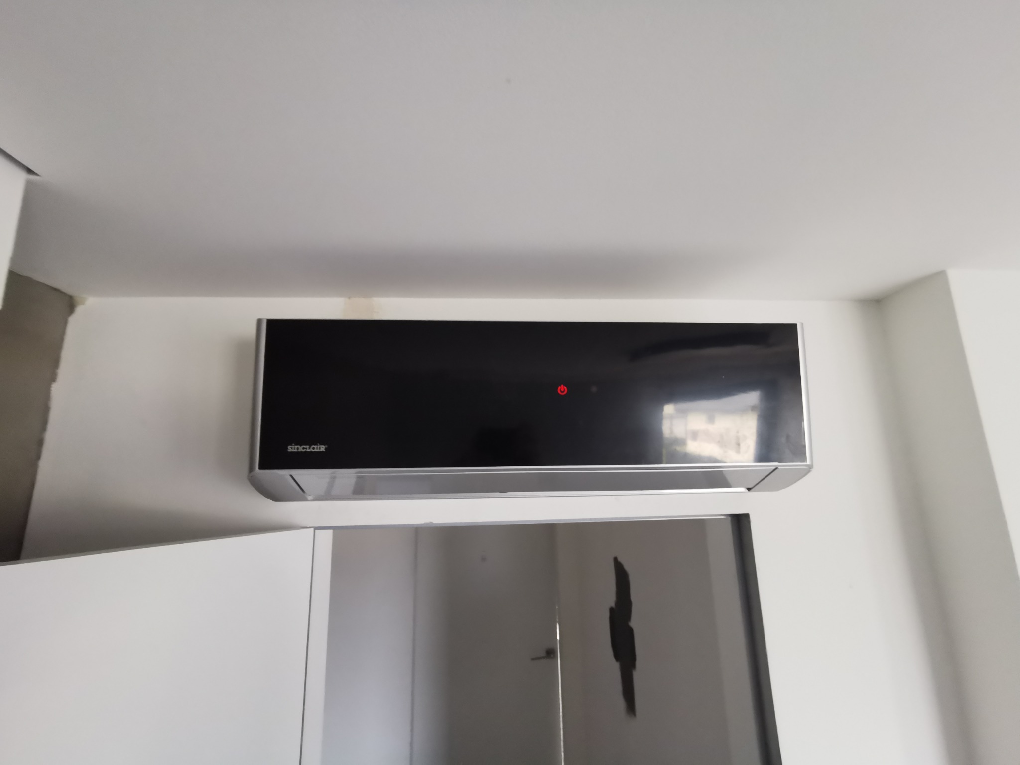 Czarny klimatyzator marki Sinclair zamontowany na białej ścianie w pomieszczeniu z białymi meblami, widoczny czerwony wskaźnik pracy urządzenia.