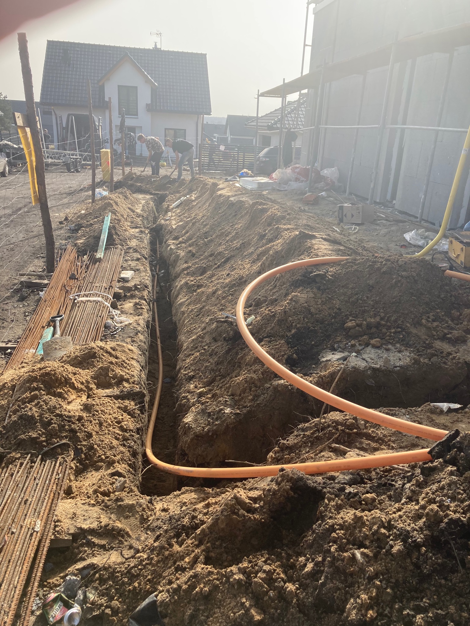 Wykop pod instalację kabla elektrycznego na budowie domu jednorodzinnego, widoczni pracownicy z łopatami, pomarańczowy kabel w wykopie, przygotowanie do podłączenia.