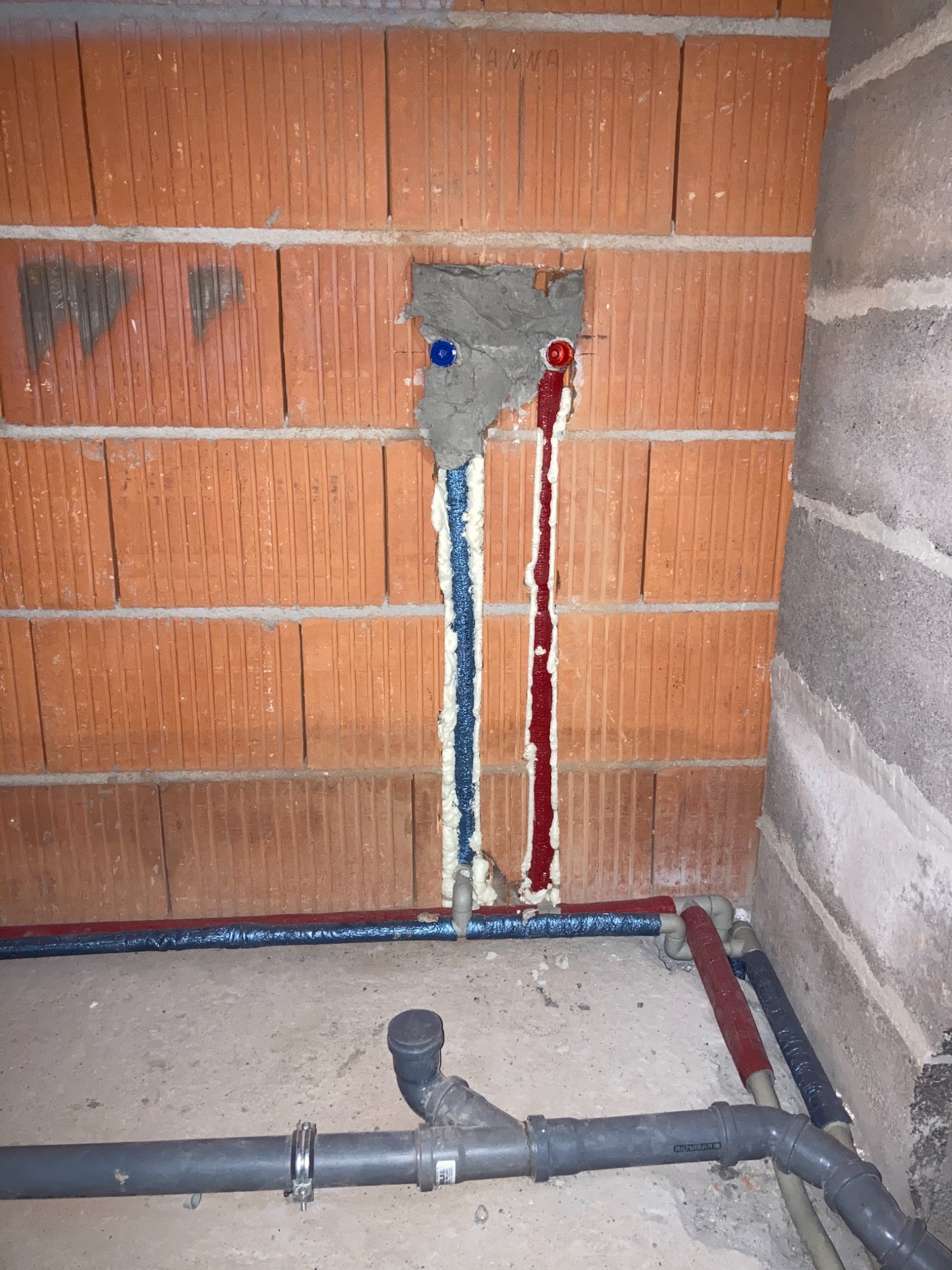 Wewnętrzna instalacja wodno-kanalizacyjna w surowym stanie: widoczne rury, izolacja termiczna w kolorach niebieskim i czerwonym, oraz fragment ściany z cegły z widocznymi otworami na przyłącza...