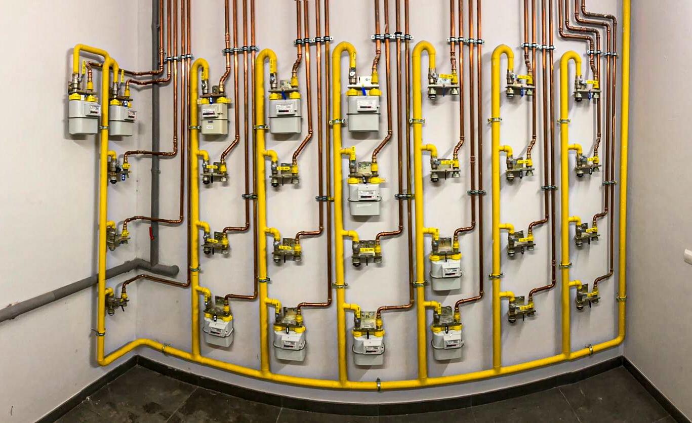 Równoległe piony instalacji gazowej z licznikami, rury miedziane i żółte, zamocowane do ściany w pomieszczeniu.