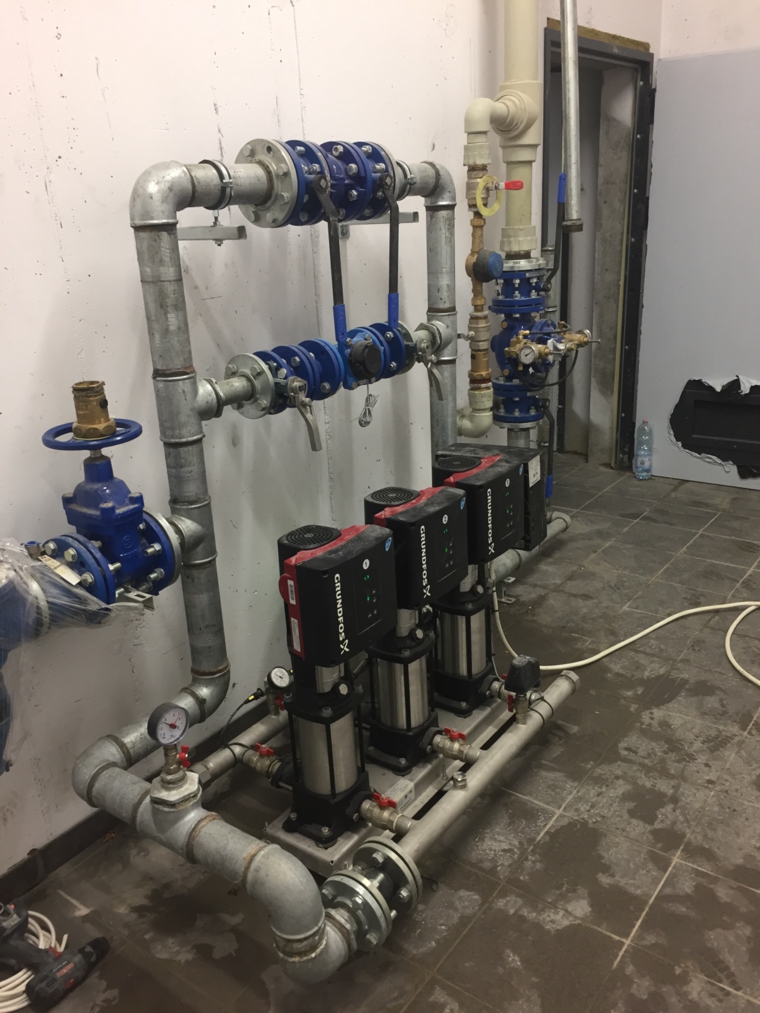 Instalacja pomp Grundfos X z rurami i zaworami w pomieszczeniu technicznym, widoczne manometry i narzędzia w tle.