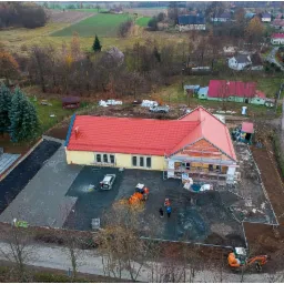 Widok z drona na plac budowy z częściowo ukończonym budynkiem z czerwonym dachem, przygotowanym podłożem pod kostkę brukową z widoczną warstwą geowłókniny, oraz pracującymi maszynami i robotnikami.