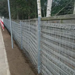 Nowo postawione, ocynkowane ogrodzenie panelowe na granicy chodnika rowerowego i drewnianego płotu, z widocznymi słupkami i świeżo wyr&oacute;wnaną ziemią u podstawy.