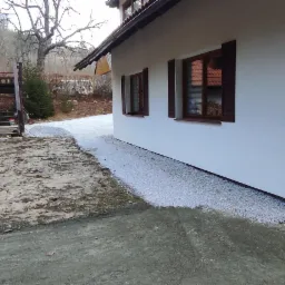 Przygotowany teren pod przyszłą ścieżkę z wysypanym drobnym białym żwirem obok białego domu z brązowymi okiennicami, widoczny teren ziemny przed domem i stertą drewna.