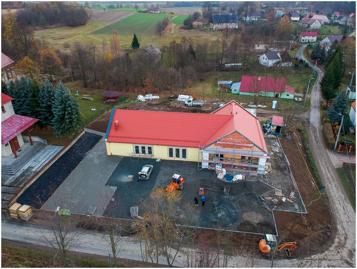 Widok z drona na plac budowy z częściowo ukończonym budynkiem z czerwonym dachem, przygotowanym podłożem pod kostkę brukową z widoczną warstwą geowłókniny, oraz pracującymi maszynami i robotnikami.