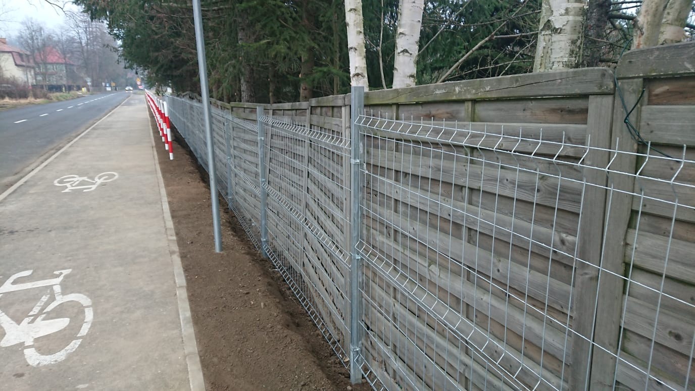 Nowo postawione, ocynkowane ogrodzenie panelowe na granicy chodnika rowerowego i drewnianego płotu, z widocznymi słupkami i świeżo wyrównaną ziemią u podstawy.