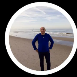 Mężczyzna w niebieskiej bluzie i ciemnych spodniach stoi na piaszczystej plaży z falami morskimi i lecącymi mewami w tle, całość kadrowana w okrągłej ramie.