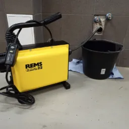 Żółta maszyna REMS Cobra 22 do czyszczenia rur, z wężem włożonym do odpływu w ścianie obok czarnego wiadra na szmatce.