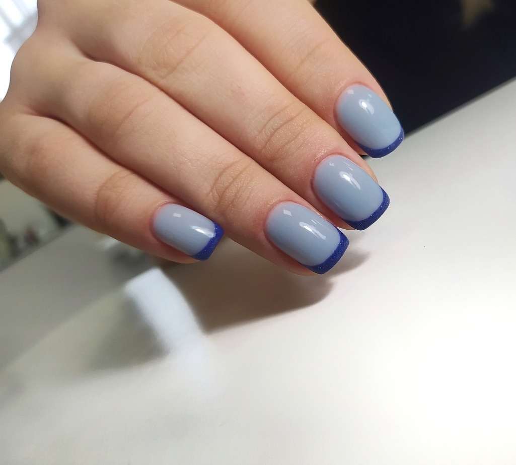 Dłoń z błękitnymi paznokciami w stylu french z granatową końcówką i delikatnym połyskiem, ułożona na białym tle.