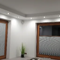 Nowoczesne wnętrze z drewnianą stolarką okienną i szarymi plisami, oświetlone punktowymi lampami sufitowymi i wyposażone w system alarmowy z czujnikiem na ścianie.