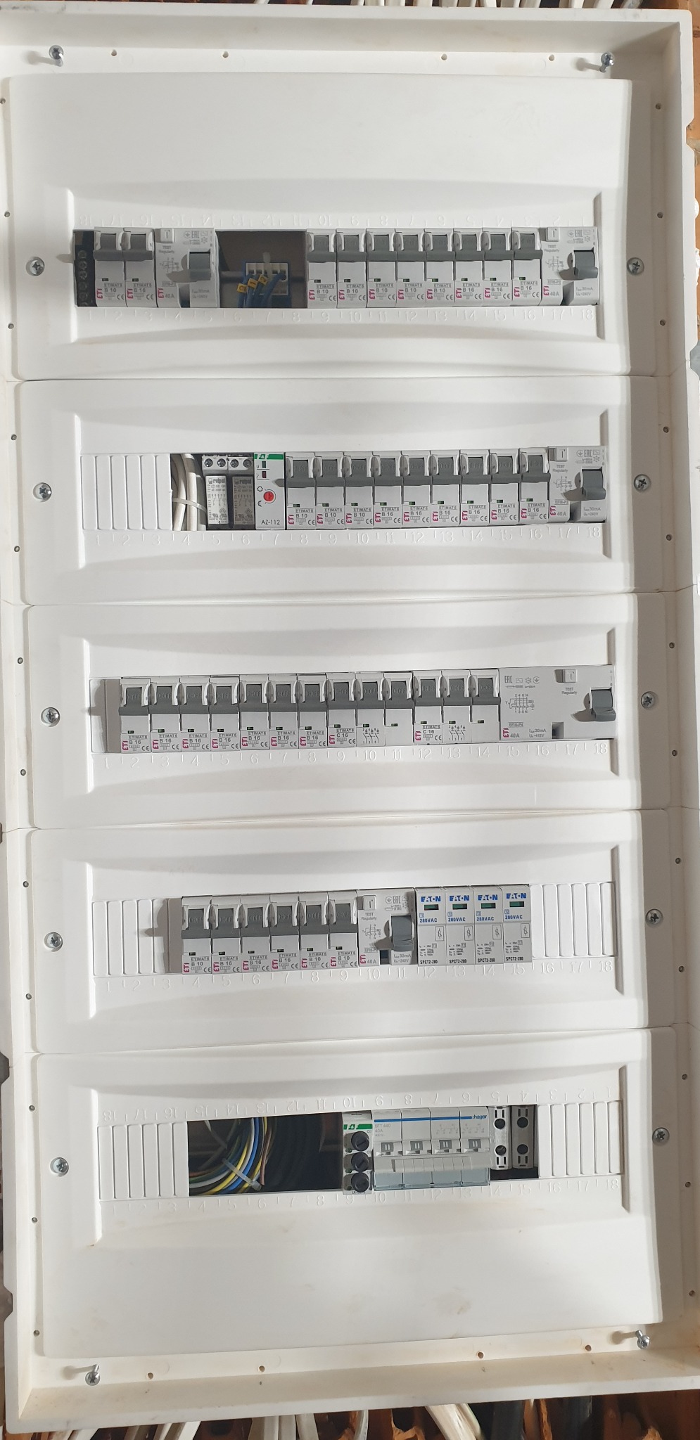 Rozdzielnica elektryczna z widocznymi bezpiecznikami i przewodami, zamontowana w białej obudowie, z numeracją obwodów od 1 do 18, widoczne różne typy zabezpieczeń.