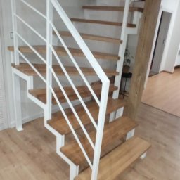 Hard Stal - Nowoczesne schody wewnętrzne z drewnianymi stopniami i białą, metalową balustradą, widoczne z dołu, z elementami drewnianej kolumny w tle.