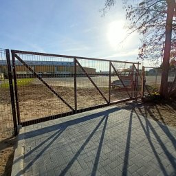 Hard Stal - Zamknięta, brązowa brama przemysłowa z metalowej siatki, z widocznym budynkiem w tle, oświetlona silnym słońcem rzucającym długie cienie na brukowaną nawierzchnię.