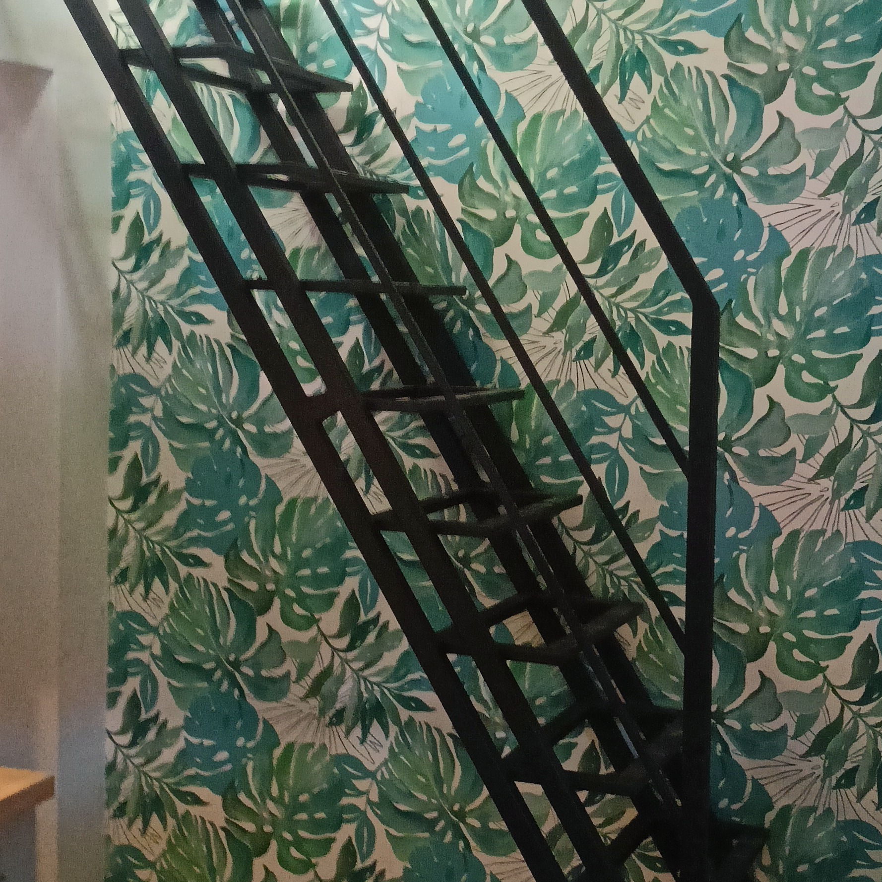 Czarne, metalowe schody o prostej konstrukcji na tle ściany z tapetą w duże, zielone liście monstery.