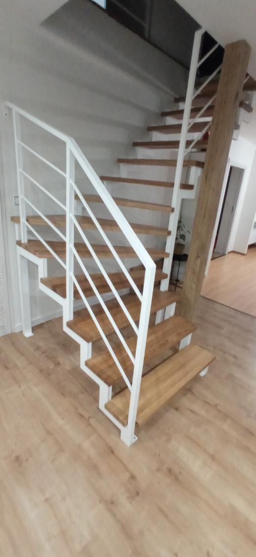 Nowoczesne schody wewnętrzne z drewnianymi stopniami i białą, metalową balustradą, widoczne z dołu, z elementami drewnianej kolumny w tle.