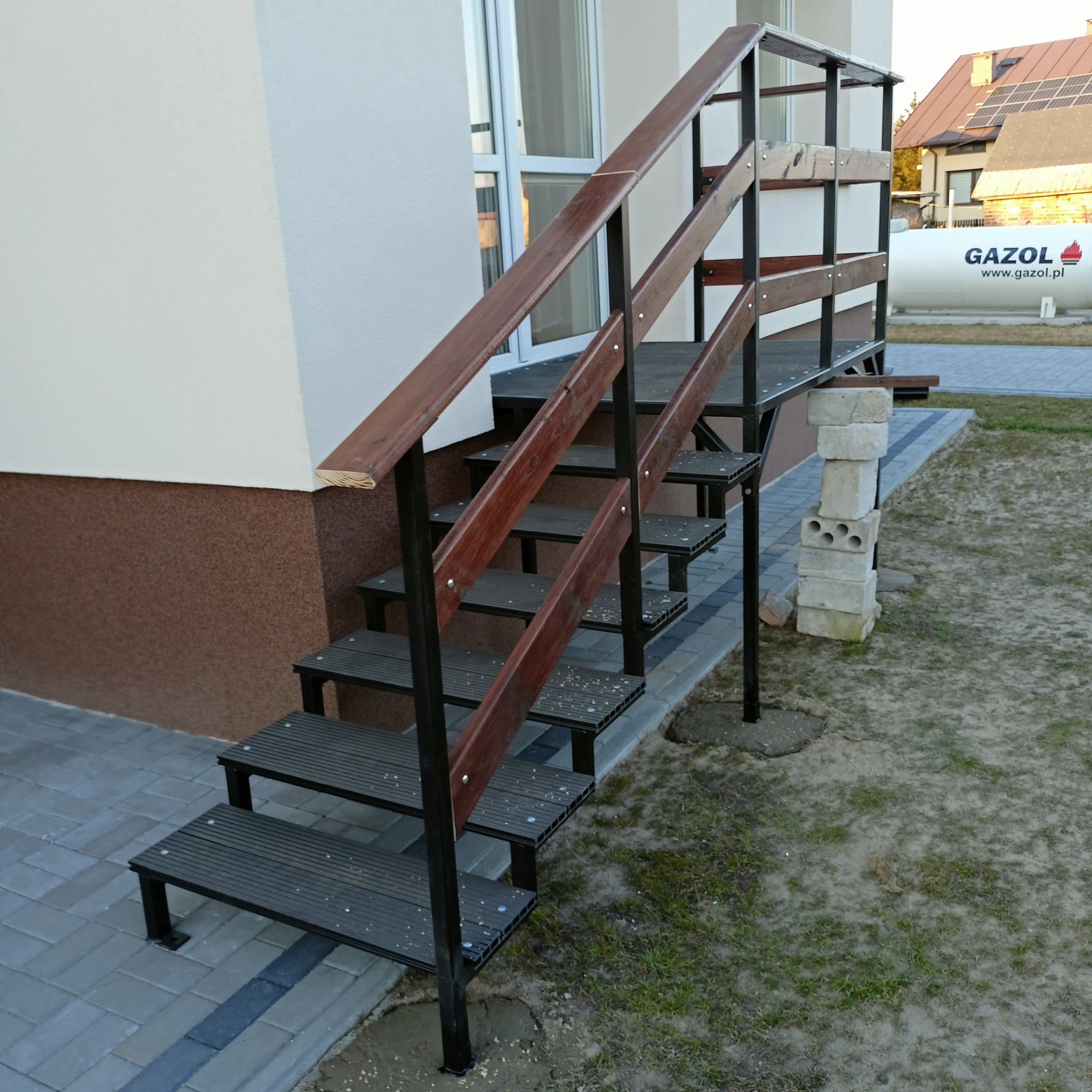 Zewnętrzne schody metalowe z drewnianymi poręczami, prowadzące do drzwi wejściowych budynku, z widoczną instalacją gazową w tle.