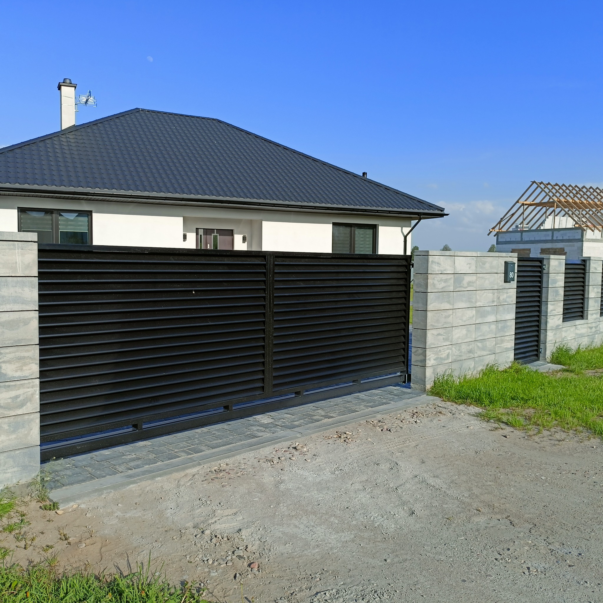 Nowoczesna, przesuwna brama wjazdowa z czarnymi, poziomymi panelami, osadzona między betonowymi słupkami, prowadząca do domu z szarą dachówką i widoczną konstrukcją drugiego budynku w tle, na tle...