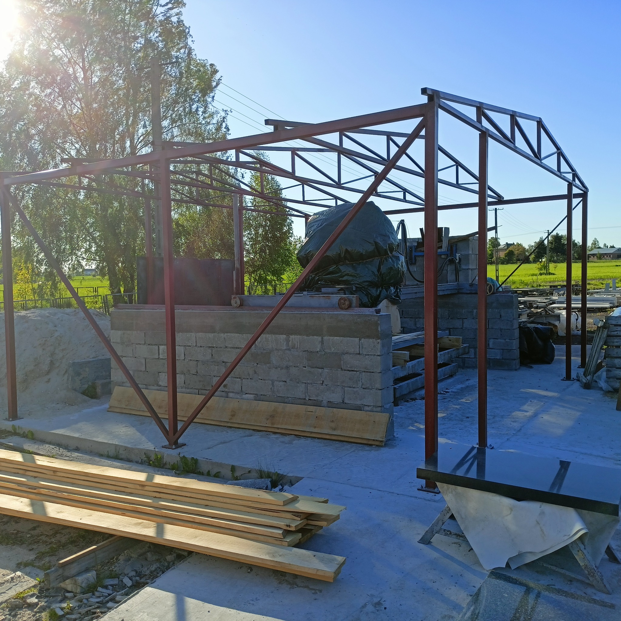 Wznoszona konstrukcja z metalowych profili na betonowej podstawie, prawdopodobnie zadaszenie nad stanowiskiem pracy z maszyną przykrytą brezentem, w tle zielone pola i drzewa.