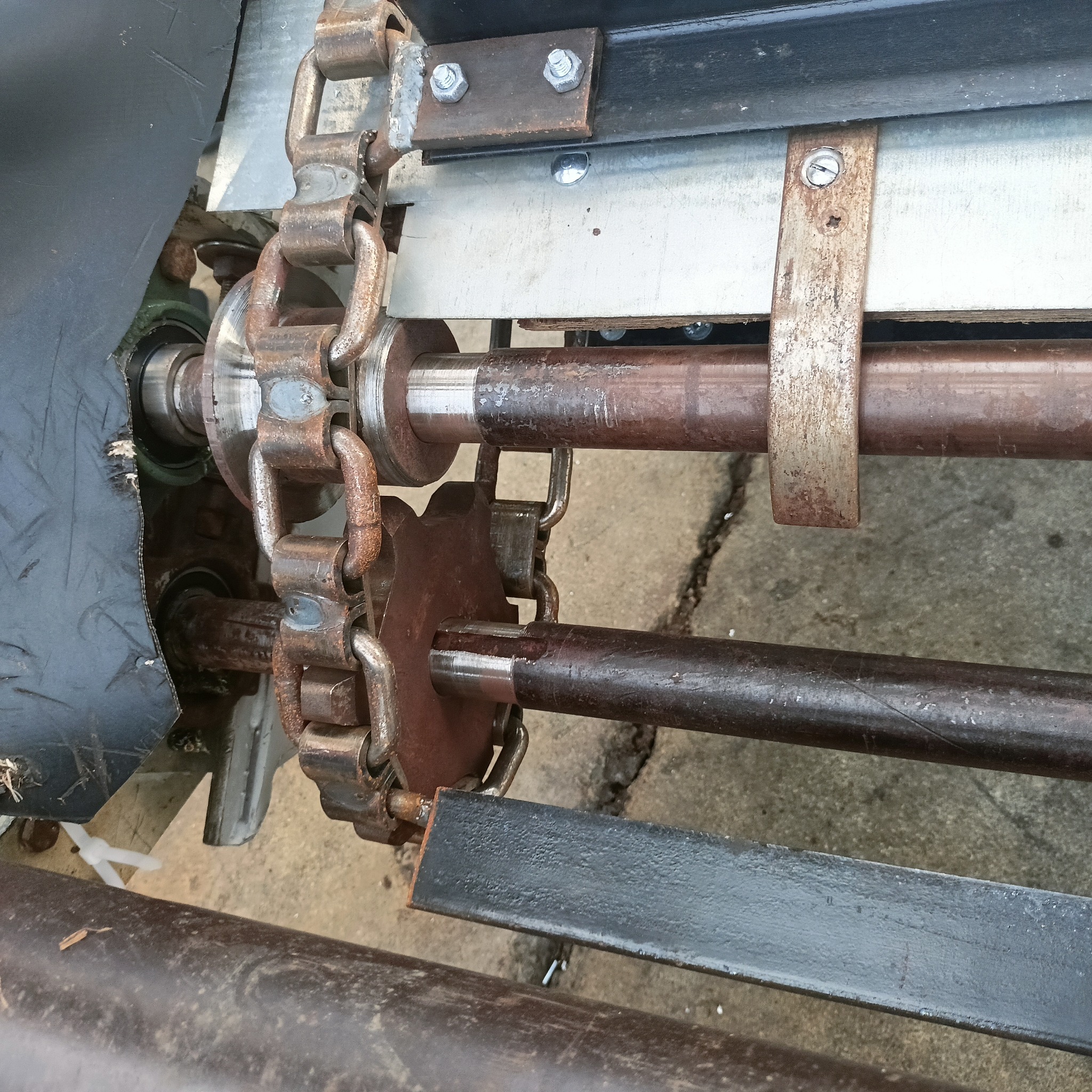 Fragment mechanizmu z łańcuchem i zębatkami, widoczne ślady korozji na metalowych elementach, konstrukcja z profili i rur, połączona śrubami.