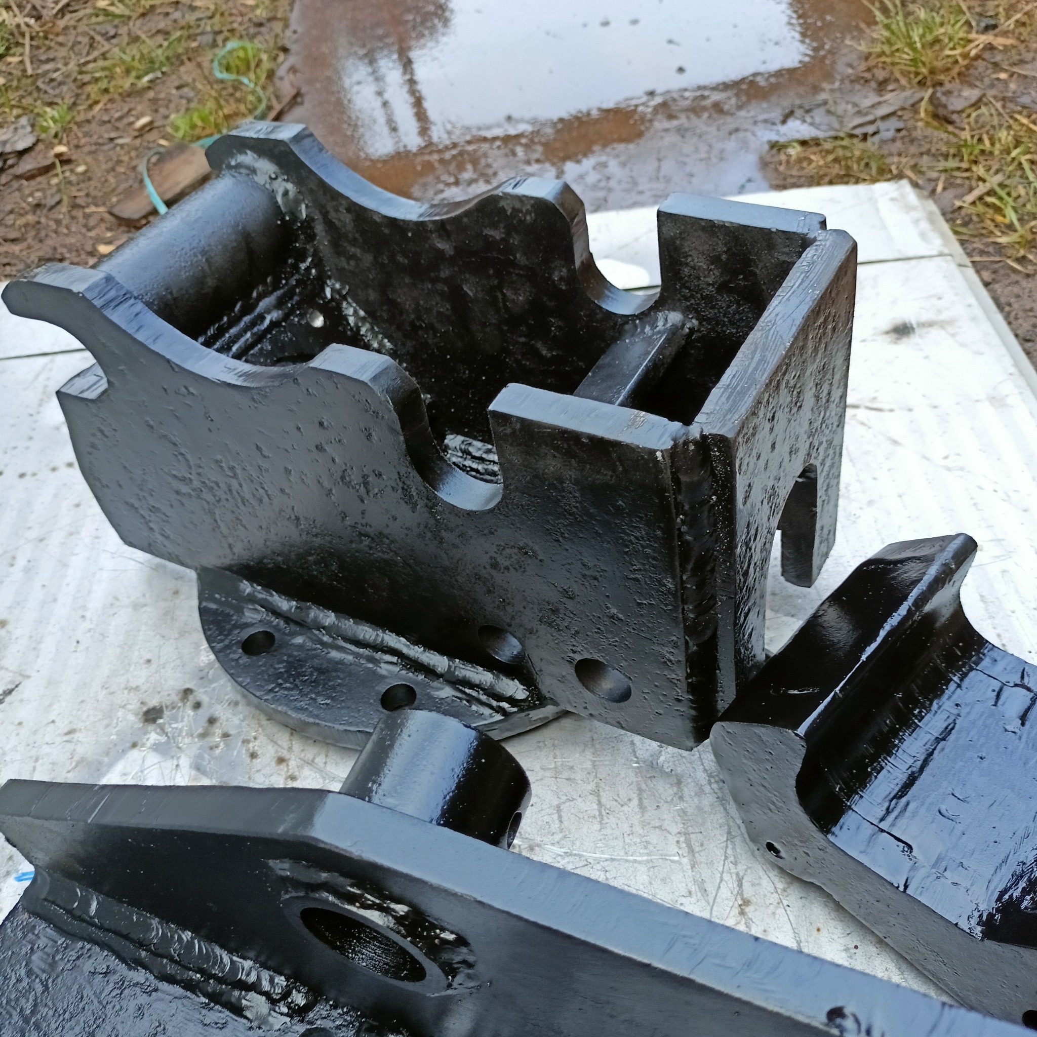 Czarne, pomalowane elementy metalowe o nieregularnych kształtach, prawdopodobnie po obróbce, leżące na szarej powierzchni na zewnątrz.