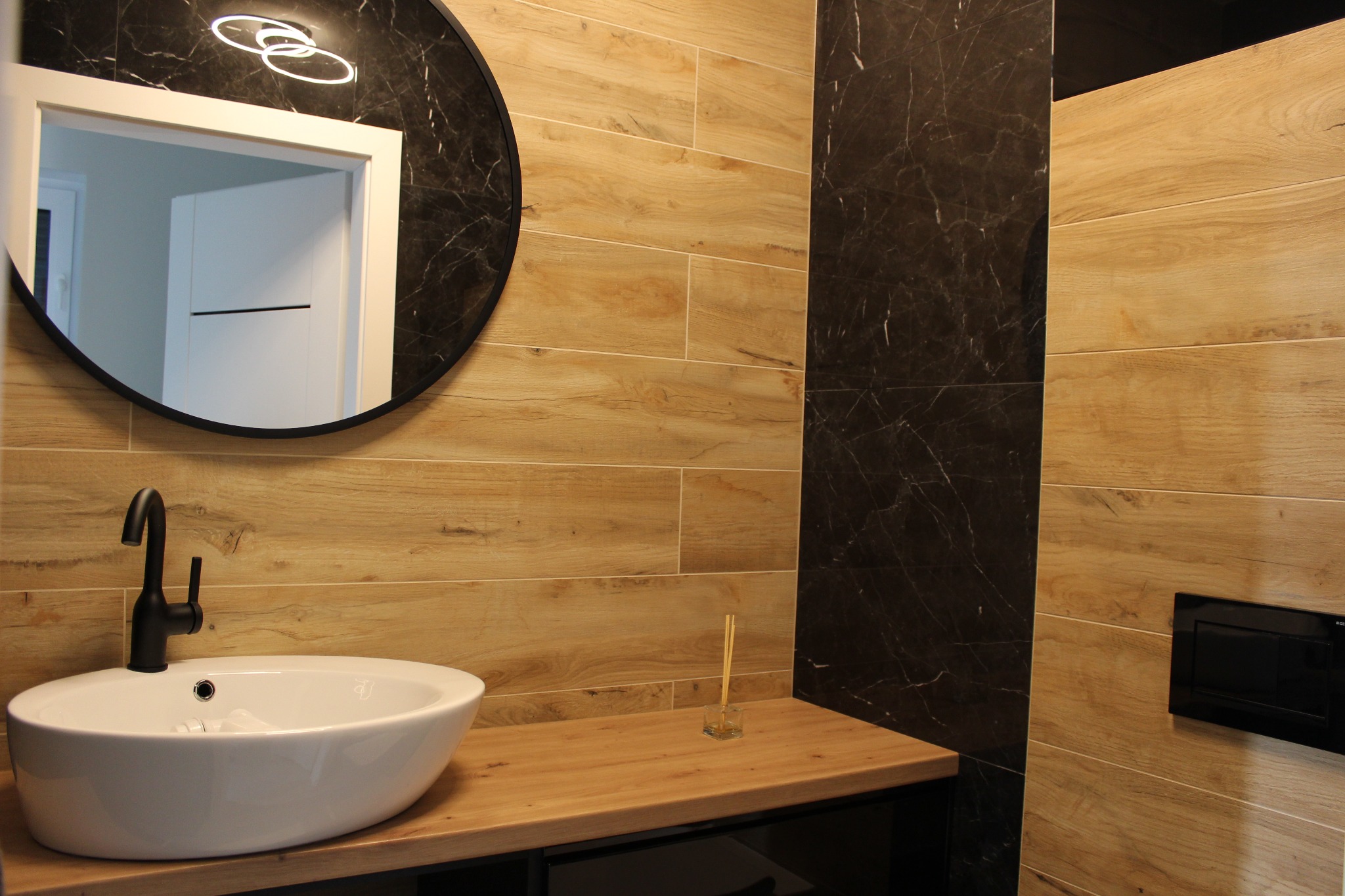 Nowoczesna łazienka z drewnopodobnymi płytkami, okrągłym lustrem z podświetleniem LED, białą umywalką nablatową i czarną armaturą, uzupełniona o czarny panel spłuczki i zapach do wnętrz z patyczkami.