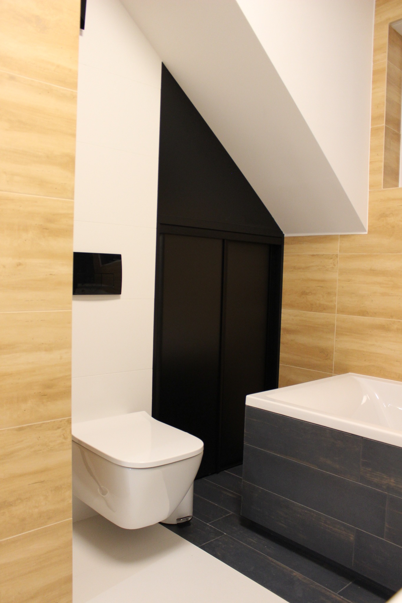 Nowoczesna łazienka z podwieszaną toaletą, obudowaną wanną w ciemnych płytkach imitujących drewno i beżowymi płytkami na ścianach, uzupełniona o czarne, przesuwne drzwi do wnęki.