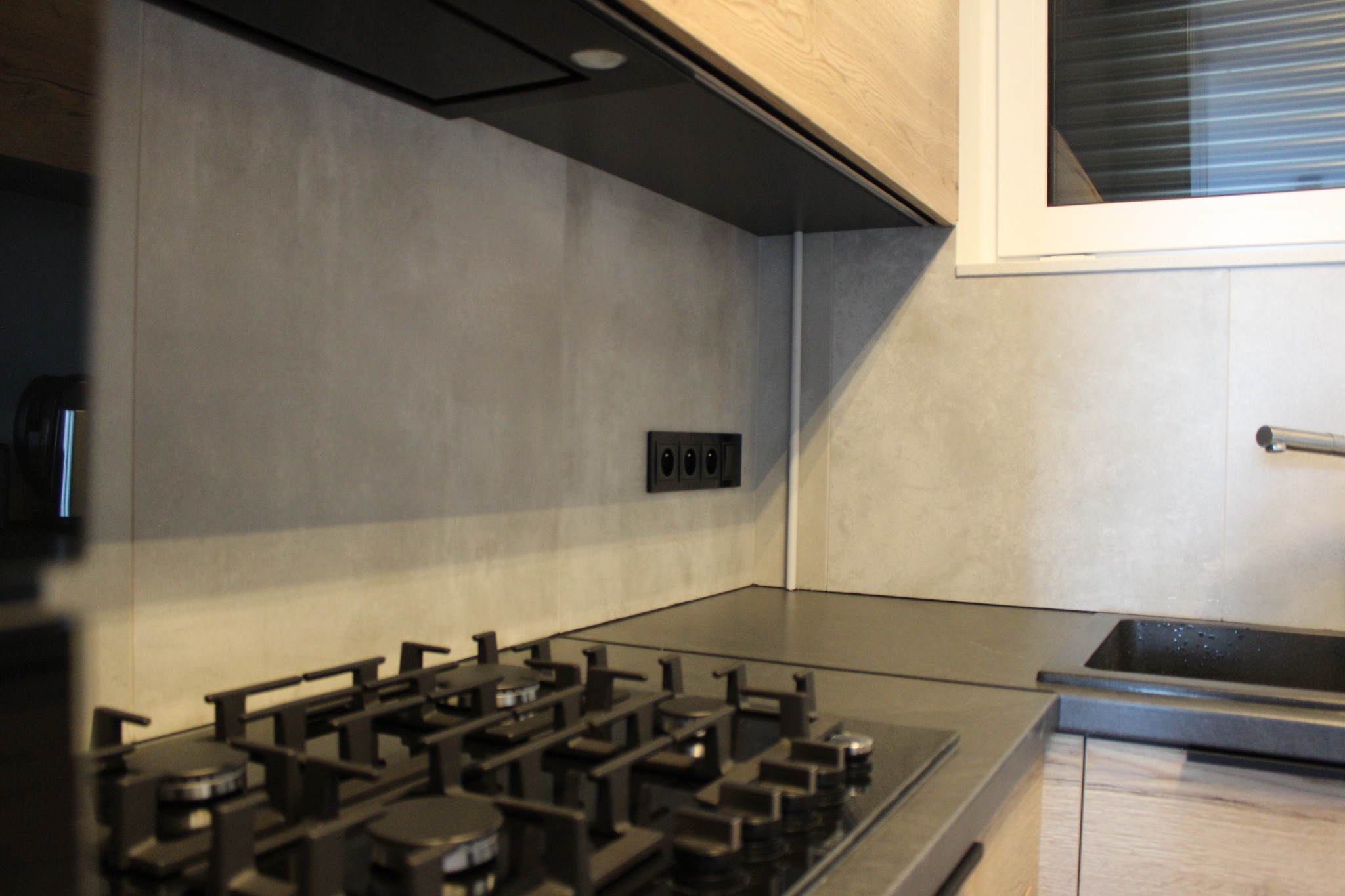 Nowoczesna kuchnia z płytą gazową, szarymi płytkami ściennymi i drewnopodobnymi szafkami, widoczne czarne gniazdka elektryczne.