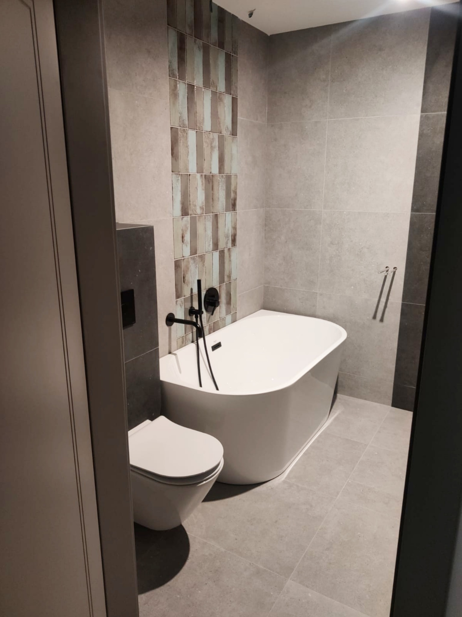 Nowoczesna łazienka z wolnostojącą białą wanną, toaletą wiszącą i szarą płytką podłogową oraz ścienną, akcentowana ścianą z dekoracyjną mozaiką w odcieniach beżu i błękitu, z czarną armaturą.