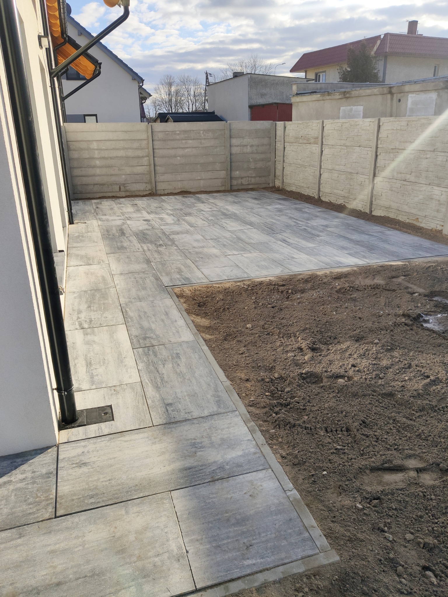 Ułożona szara kostka brukowa na podwórku z fragmentem ziemi przygotowanej pod trawnik, widoczny fragment domu z rynną i betonowe ogrodzenie.