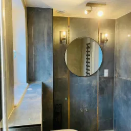 Nowoczesna łazienka z szarymi płytkami, złotymi akcentami, okrągłym lustrem i minimalistyczną toaletą.