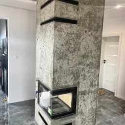 Nowoczesny kominek obudowany tynkiem strukturalnym z czarnymi, poziomymi akcentami, wkomponowany w narożnik pomieszczenia z szarą marmurową podłogą i białymi drzwiami w tle.