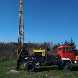 Maszyna wiertnicza z żółtym silnikiem i wysoką, metalową wieżą na podwoziu czerwonej ciężarówki, stojąca na trawiastym terenie z widocznym lasem w tle.