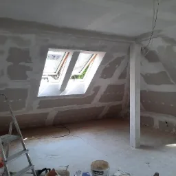 Poddasze w trakcie prac wykończeniowych, widoczne ściany z nałożoną gładzią szpachlową, okno dachowe z widokiem na zieleń oraz drabina malarska i akcesoria malarskie na podłodze.