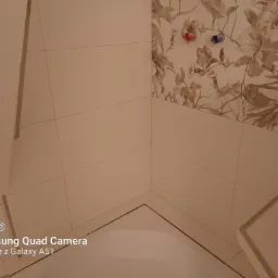 Wykończona narożna strefa w łazience z jasnymi płytkami ściennymi i dekoracyjnymi płytkami z motywem kwiatowym powyżej, widoczne przyłącza wodne.