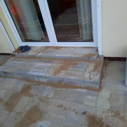 Dwu-stopniowe schody wejściowe wykonane z szarych kostek brukowych, częściowo zasypane piaskiem, prowadzące do białych drzwi balkonowych z widocznymi zasłonami w tle.