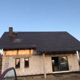 Dom w trakcie budowy z ciemnym dachem z dachówki ceramicznej, widoczne okno dachowe i komin, ściany z białego bloczka, drabina oparta o ścianę, w tle drzewa i błękitne niebo.