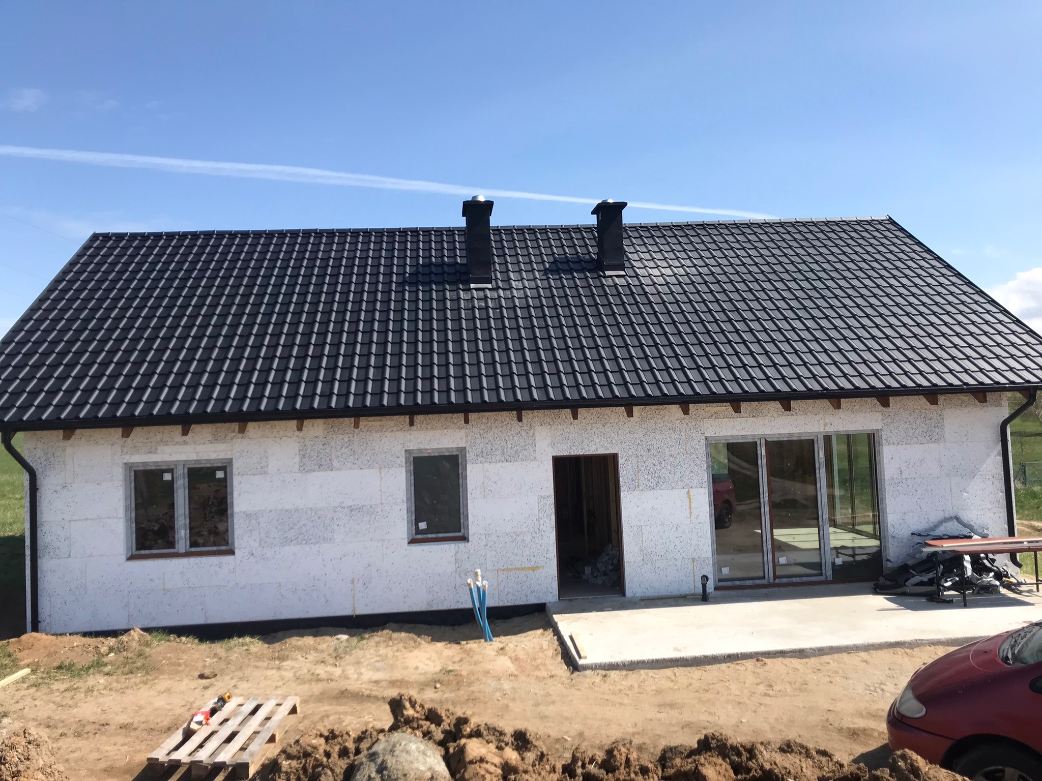 Dom w trakcie budowy z nowym, czarnym dachem z dachówki ceramicznej i dwoma kominami, widok z zewnątrz na białe ściany z oknami i betonowy wylew przed wejściem, na pierwszym planie ziemia i samochód.