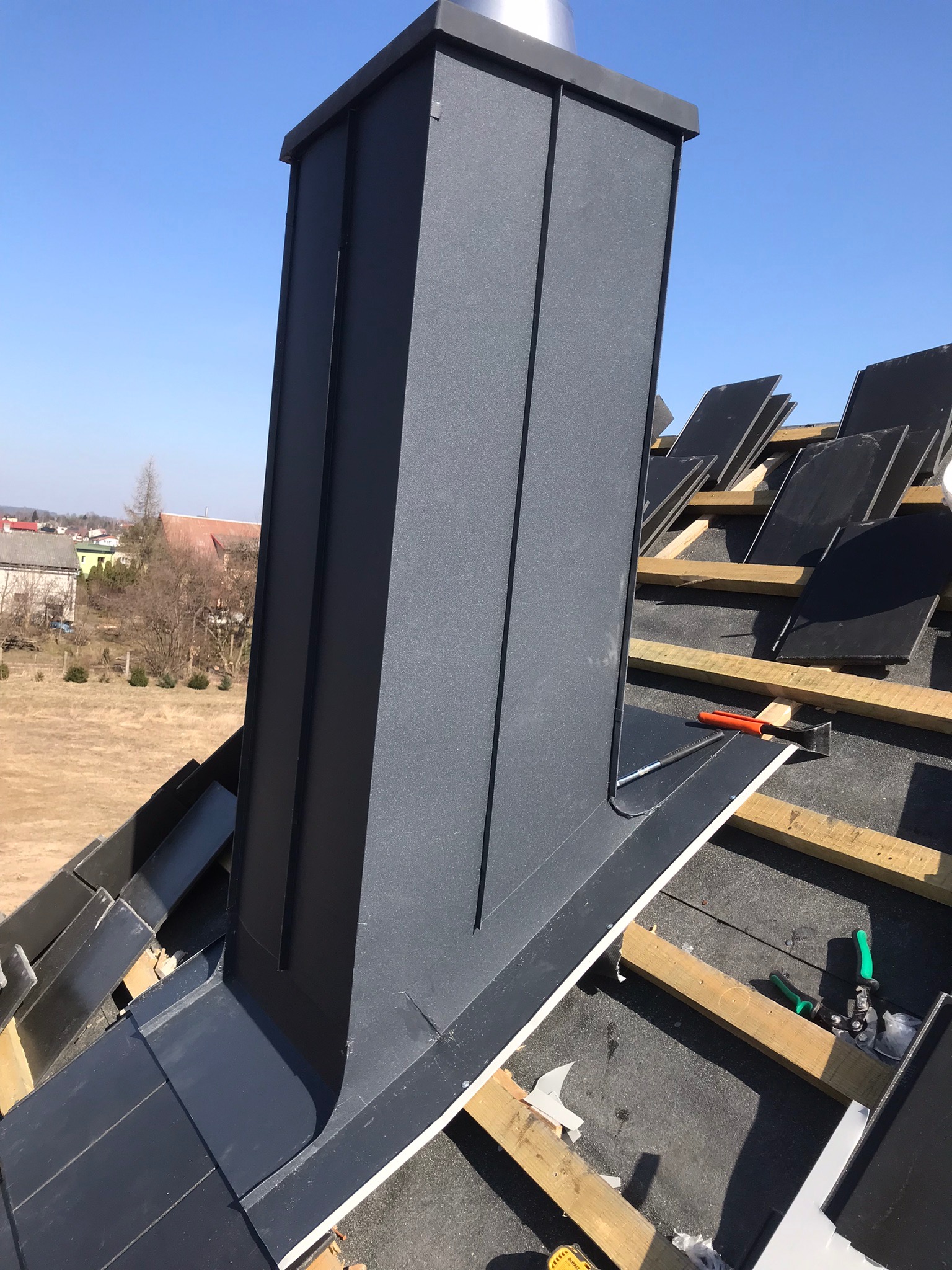 Montaż metalowego komina na dachu z częściowo ułożonymi czarnymi płytkami, widoczne narzędzia i drewniane elementy konstrukcyjne.