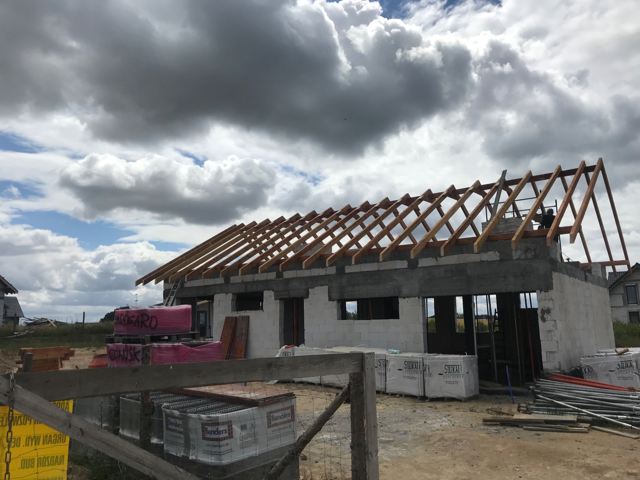 Budowa domu jednorodzinnego w stanie surowym otwartym, widoczna konstrukcja drewniana dachu, palety z dachówką i materiały budowlane na placu budowy, pochmurne niebo.