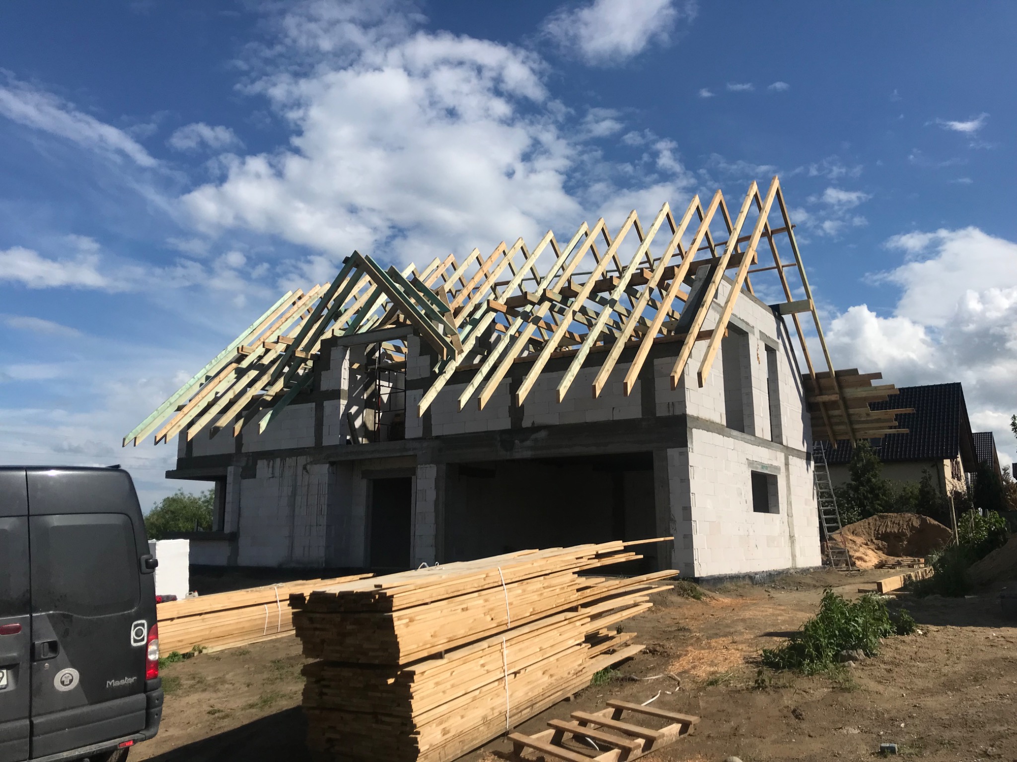 Budowa domu jednorodzinnego w trakcie montażu drewnianej konstrukcji dachu, widoczne krokwie i elementy więźby, na pierwszym planie stos desek i samochód dostawczy.