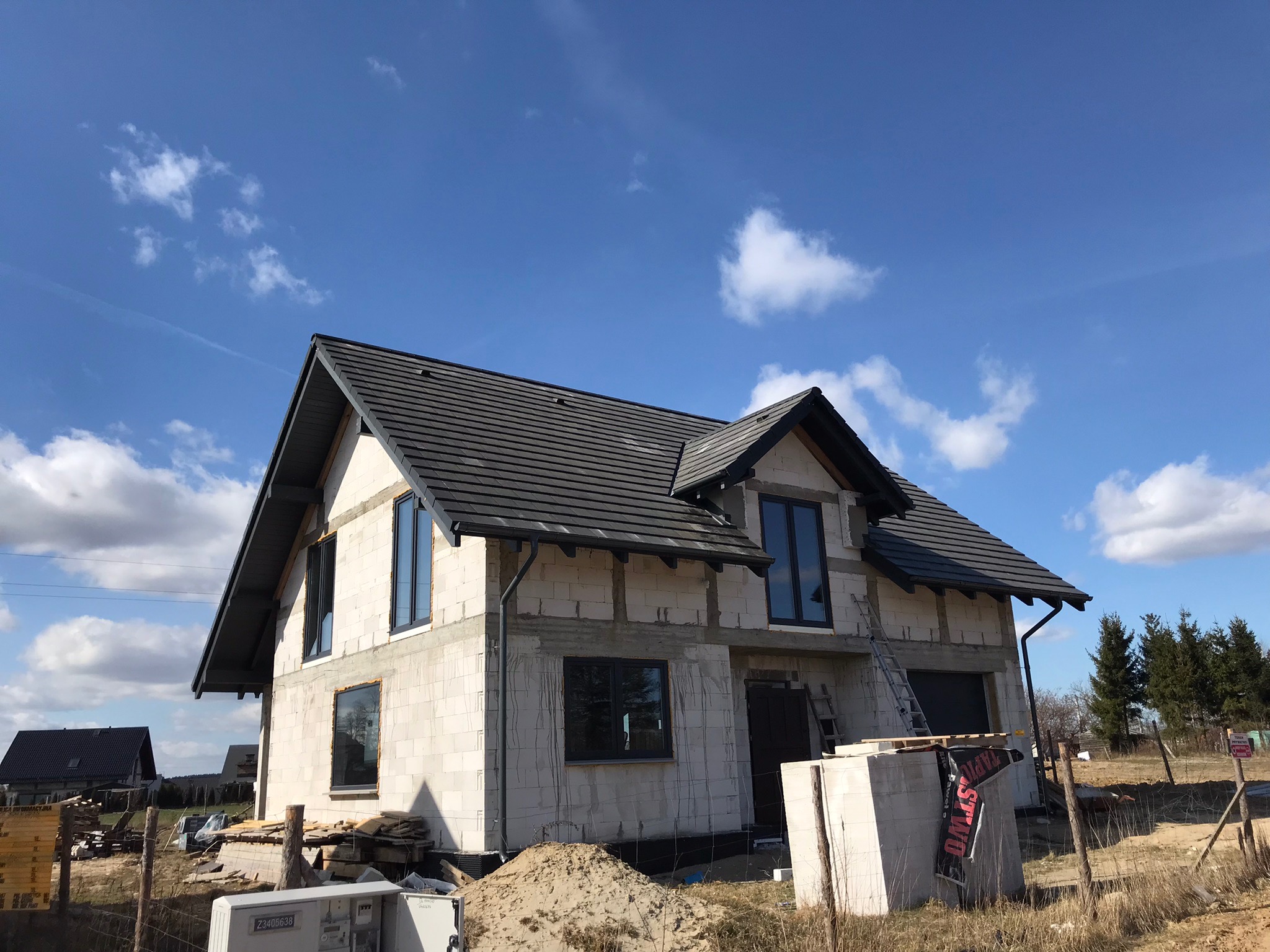 Dom w trakcie budowy z widocznym dachem z ciemnej dachówki, oknami wstawionymi w surowy mur i stertą materiałów budowlanych przed budynkiem, na tle błękitnego nieba z chmurami.