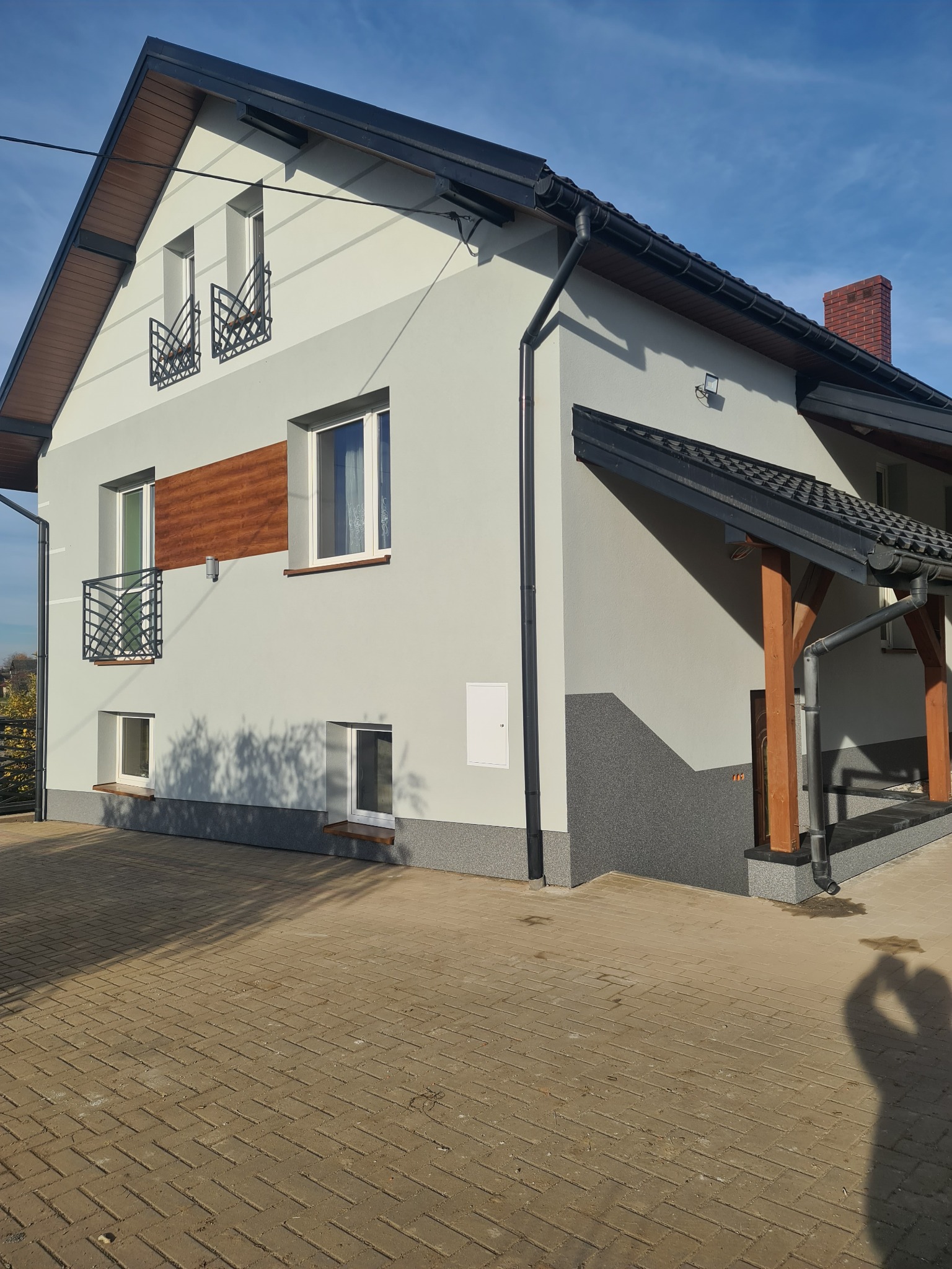 Szary dom jednorodzinny z drewnianymi akcentami i czarną dachówką, widok z zewnątrz na elewację, z brukowanym podjazdem i cieniem fotografa.