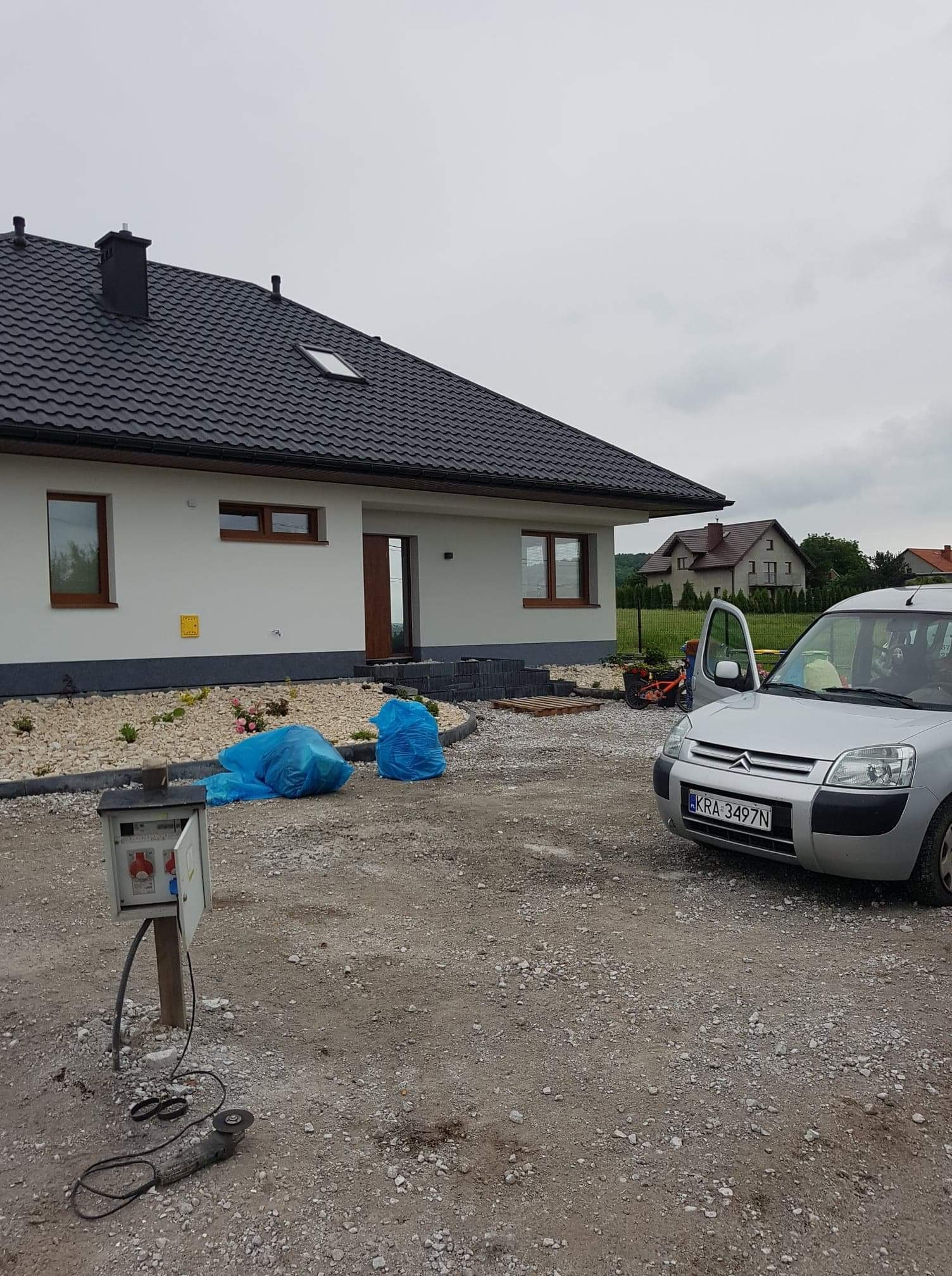 Jasny dom jednorodzinny z ciemną dachówką i brązowymi oknami, widoczne dwa worki z odpadami budowlanymi, samochód dostawczy z otwartymi drzwiami oraz skrzynka elektryczna z narzędziem do cięcia...