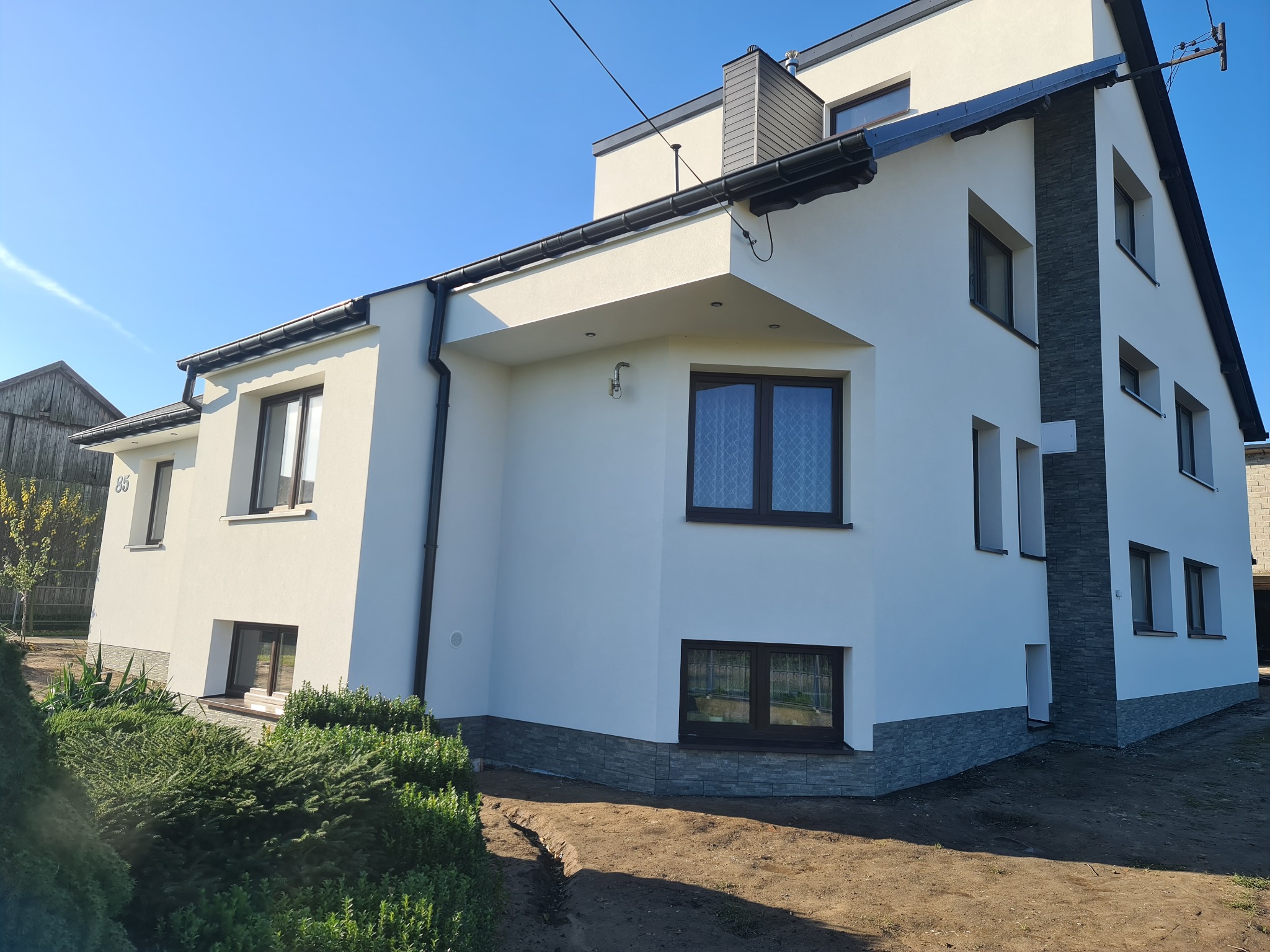 Nowoczesny dom z białą elewacją, ciemnymi oknami i szarym akcentem kamiennym, widok z zewnątrz na tle błękitnego nieba.