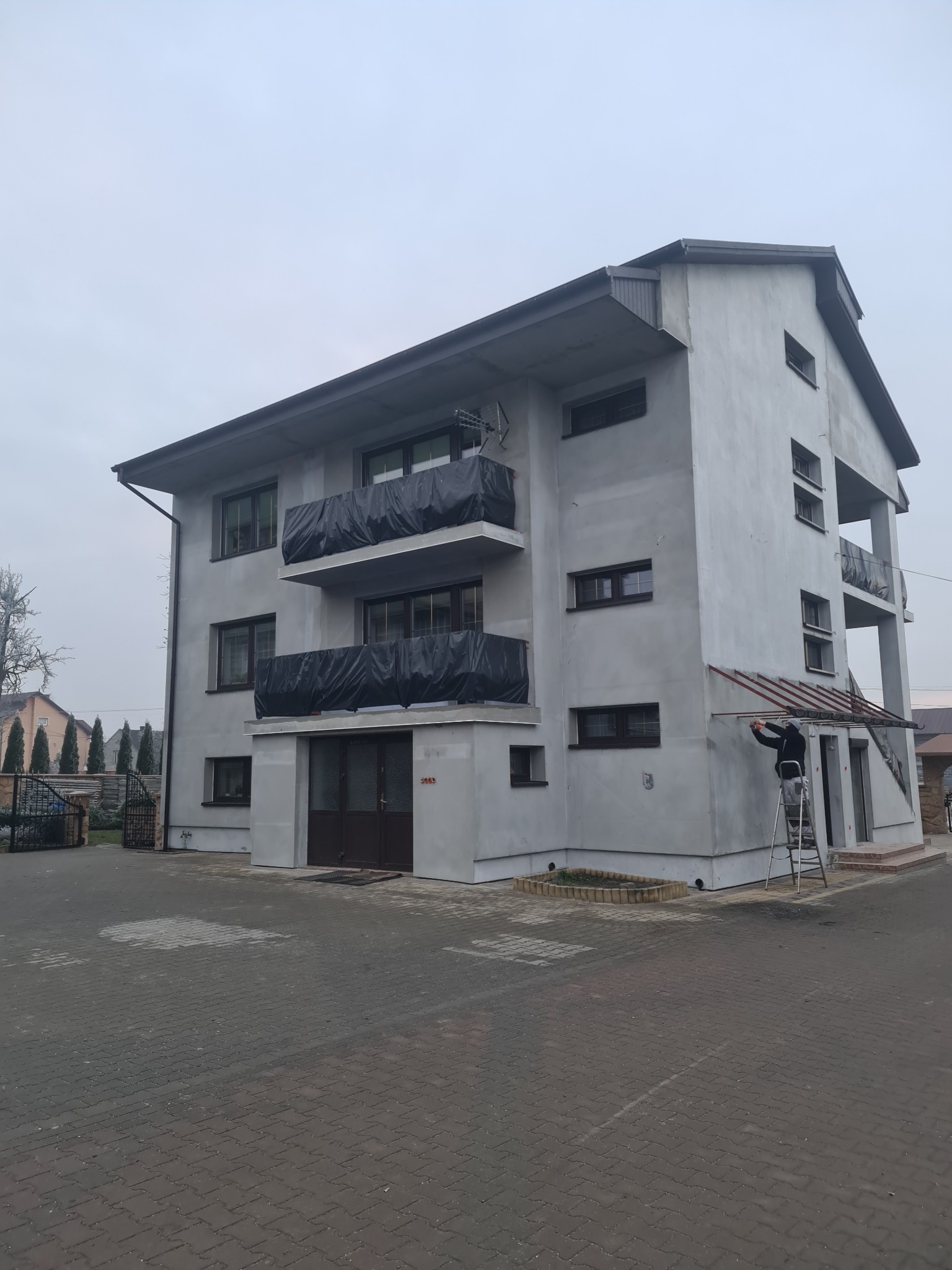 Trzykondygnacyjny budynek w trakcie prac elewacyjnych, zabezpieczone balkony folią, pracownik na drabinie przy wejściu, szara elewacja w surowym stanie, kostka brukowa na podjeździe.