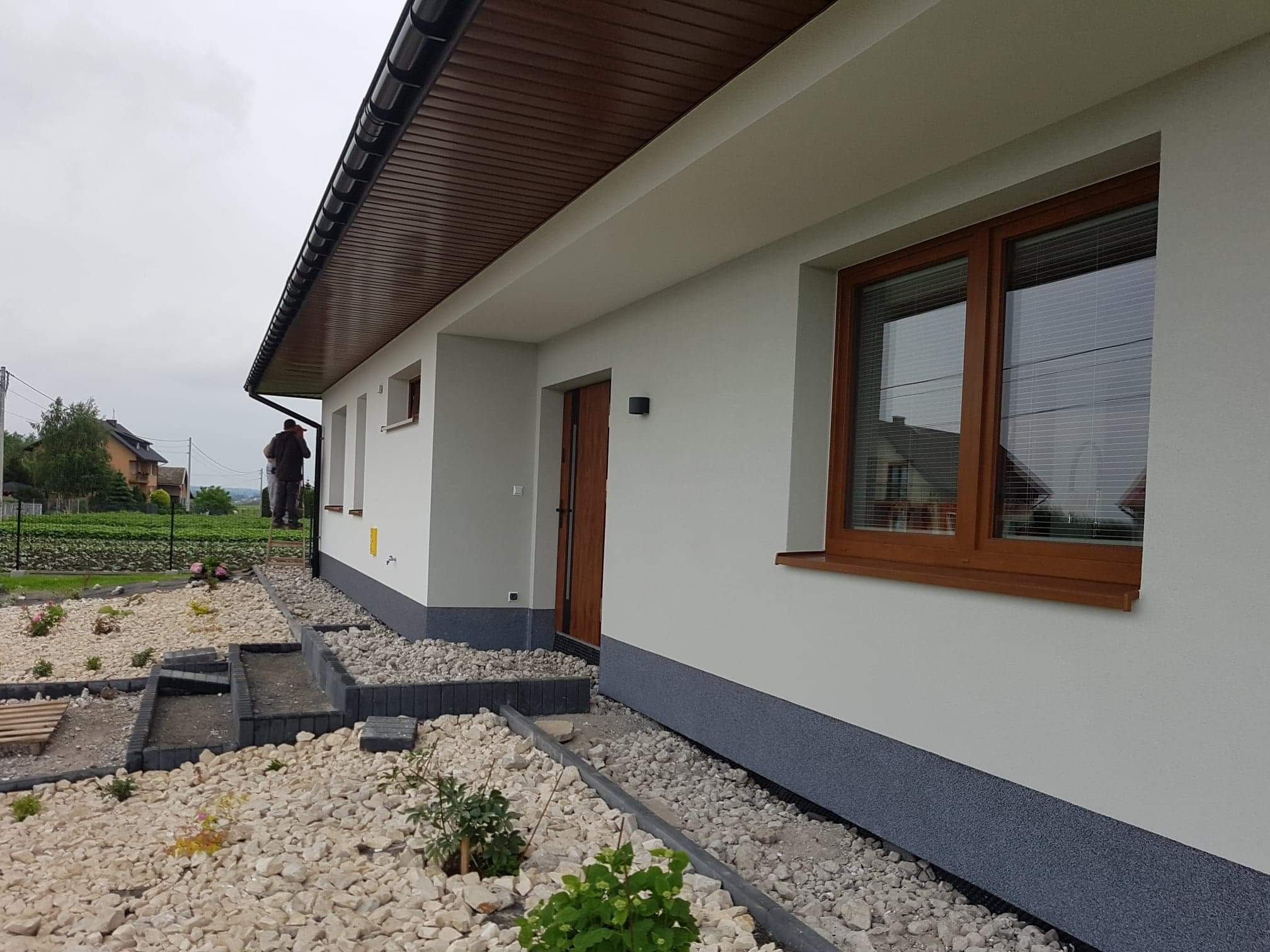 Jasnoszary dom jednorodzinny z brązowymi ramami okiennymi i drewnianym podbitką dachu, widok z boku na ogród z białym żwirem i schodkami, w tle sylwetka osoby na drabinie przy ścianie