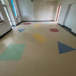 MW PODŁOGI MICHAŁ WOJCIECHOWSKI - Jasna wykładzina w pokoju z kolorowymi figurami geometrycznymi: koła, trójkąty i prostokąty. Widok na okno i grzejnik.